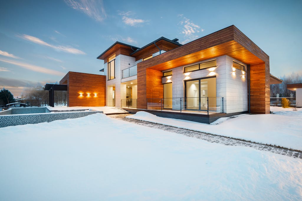 Exterior architecture of private suburban cottage house with stone and wooden facade and large windows overlooking spacious snow covered yard in winter day